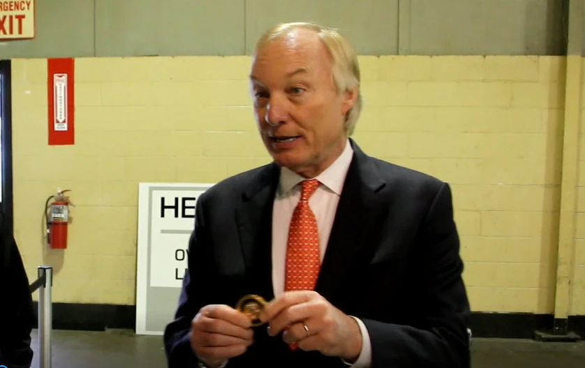 Man in dark suit and orange tie holding a small object in a hallway