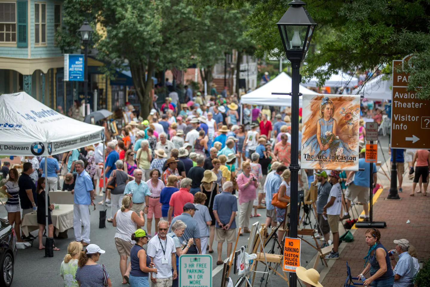 Crowded outdoor street fair with vendor tents and artwork displays on a tree-lined street