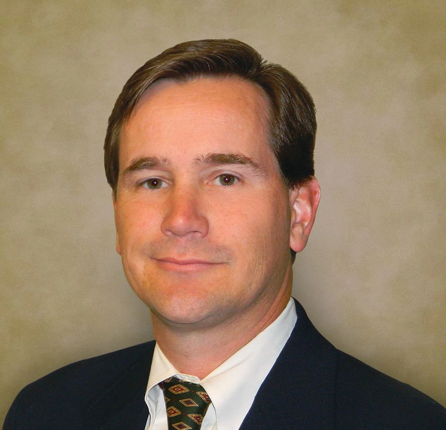 Headshot of a man in a dark suit and patterned tie against a tan backdrop