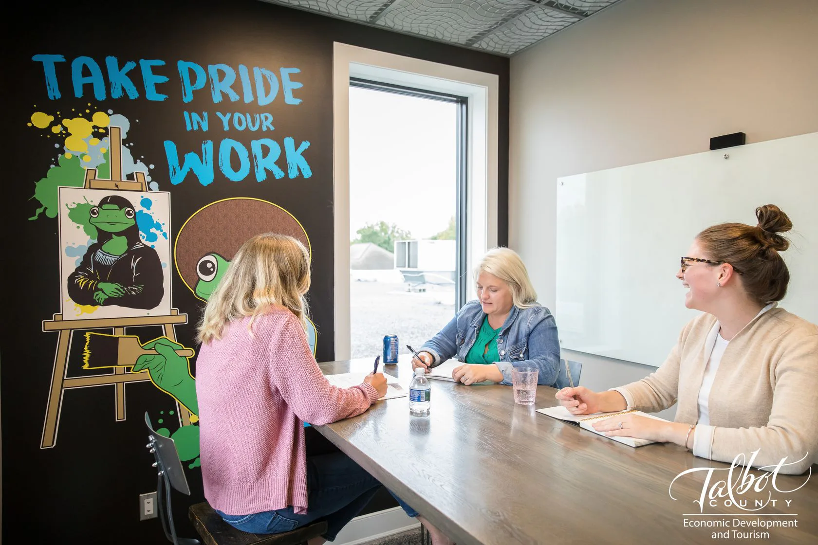 Three people meeting at a table beneath a Take Pride in Your Work wall mural