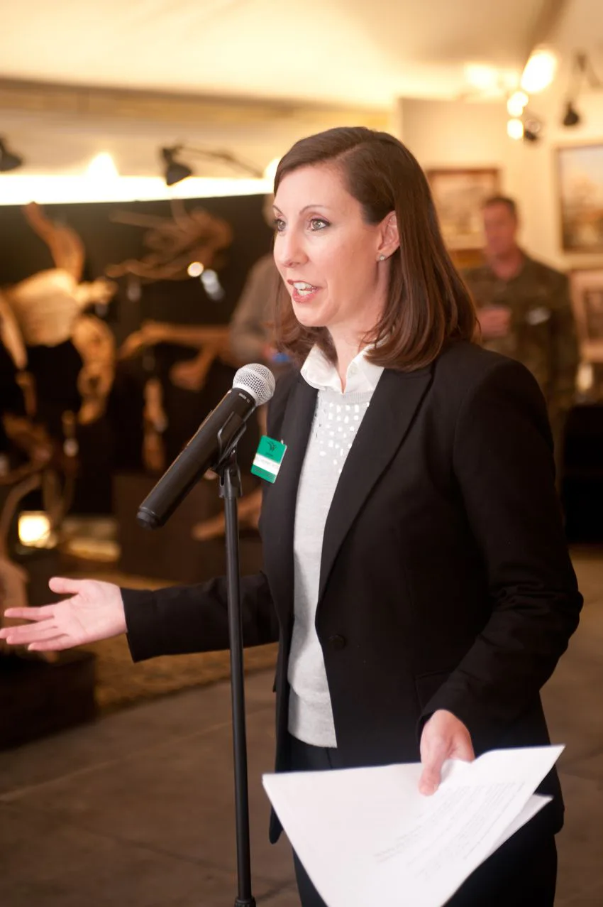 Woman in a dark blazer speaking at a microphone and reading from papers