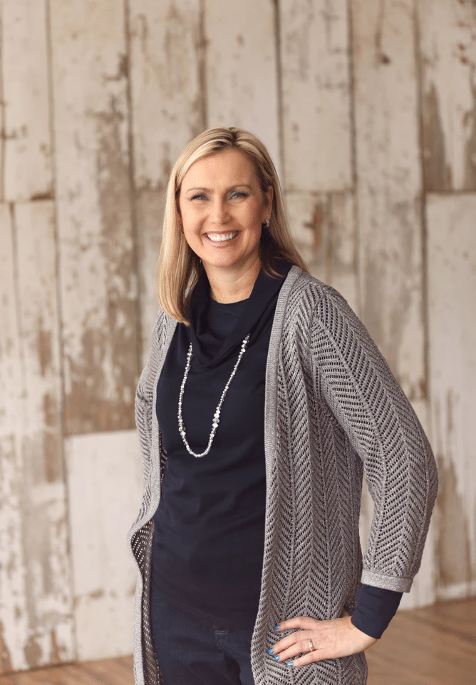 Smiling woman in a gray cardigan and necklace against a weathered wood wall