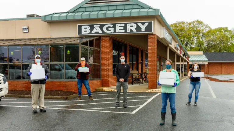 Masked volunteers holding food boxes in front of a storefront sign reading BAGERY