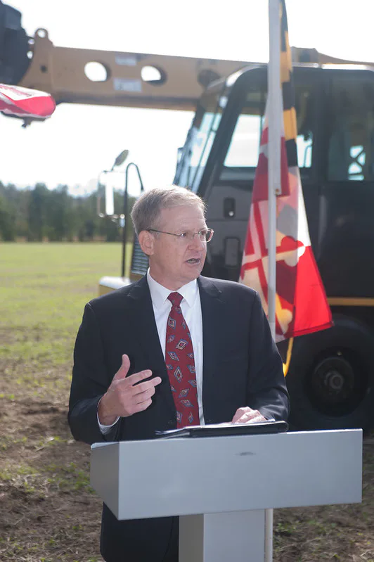 Man in a suit speaking at a podium in front of construction equipment and a flag