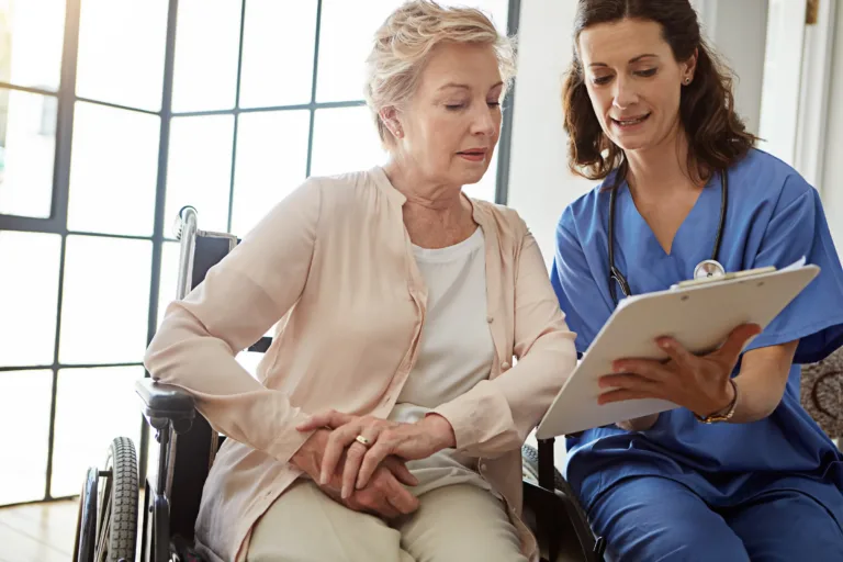 Nurse in blue scrubs reviewing results on a clipboard with an older woman in a wheelchair