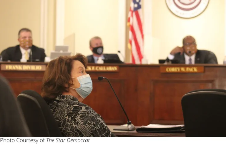 Woman wearing a face mask speaking at a county meeting, with officials seated at the dais