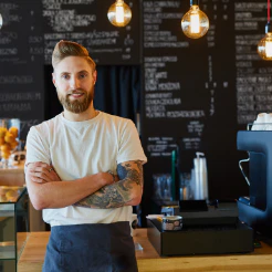 Bearded man with tattooed arms crossed in a cafe with a chalkboard menu