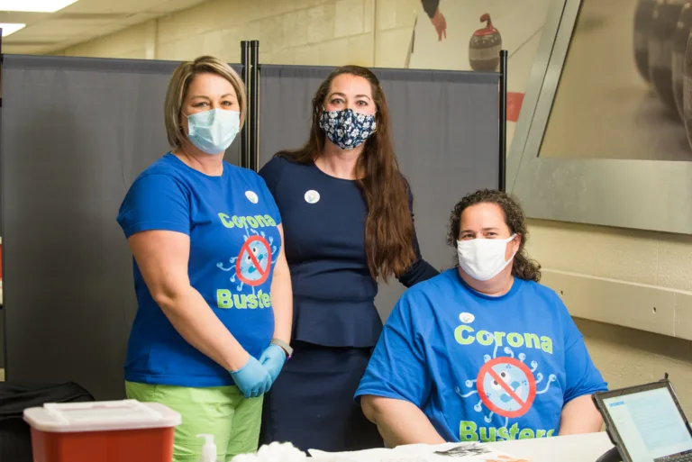 Three people in blue Corona Busters shirts at a COVID-19 vaccination station