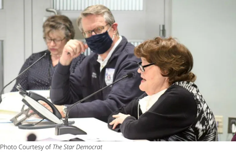 Three people seated at a meeting table, one wearing a face mask
