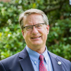 Smiling man with glasses in a suit and red tie in front of green shrubs