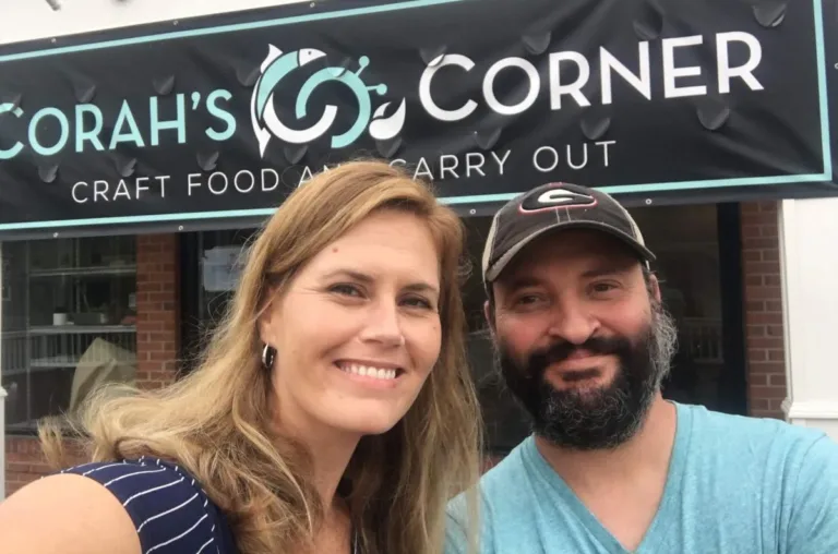 Couple smiling in front of a Corah's Corner sign reading CRAFT FOOD AND CARRY OUT