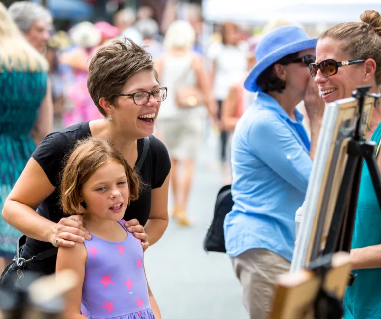 Smiling woman and young girl at an outdoor art fair with paintings nearby