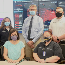 Five people posed in a control room in front of a COVID-19 dashboard screen