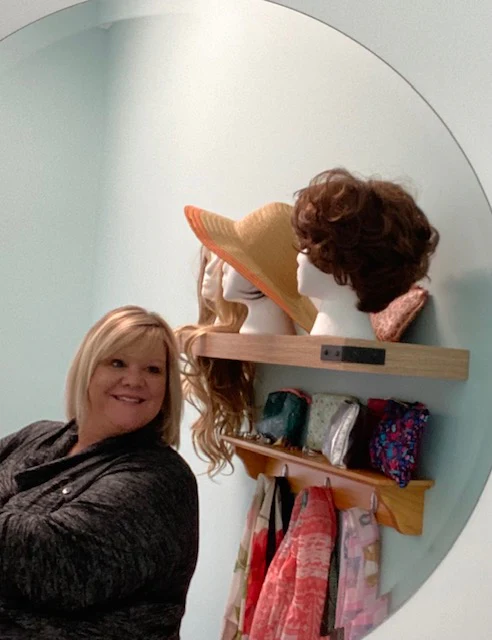 Woman smiling beside a shelf displaying wigs, a hat and scarves