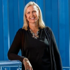 Smiling blonde woman in a black top and statement necklace in front of blue panels