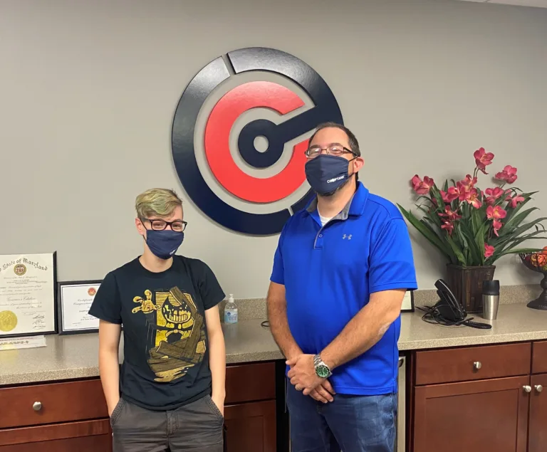 Two masked people posing in an office beneath a red and blue circular C logo