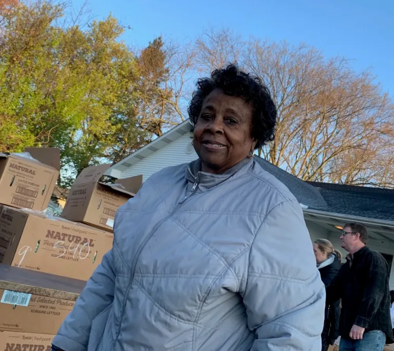 Smiling woman in a light puffer jacket beside stacked cardboard food boxes