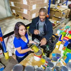 Two workers filling cans in a warehouse with shelves and boxes behind