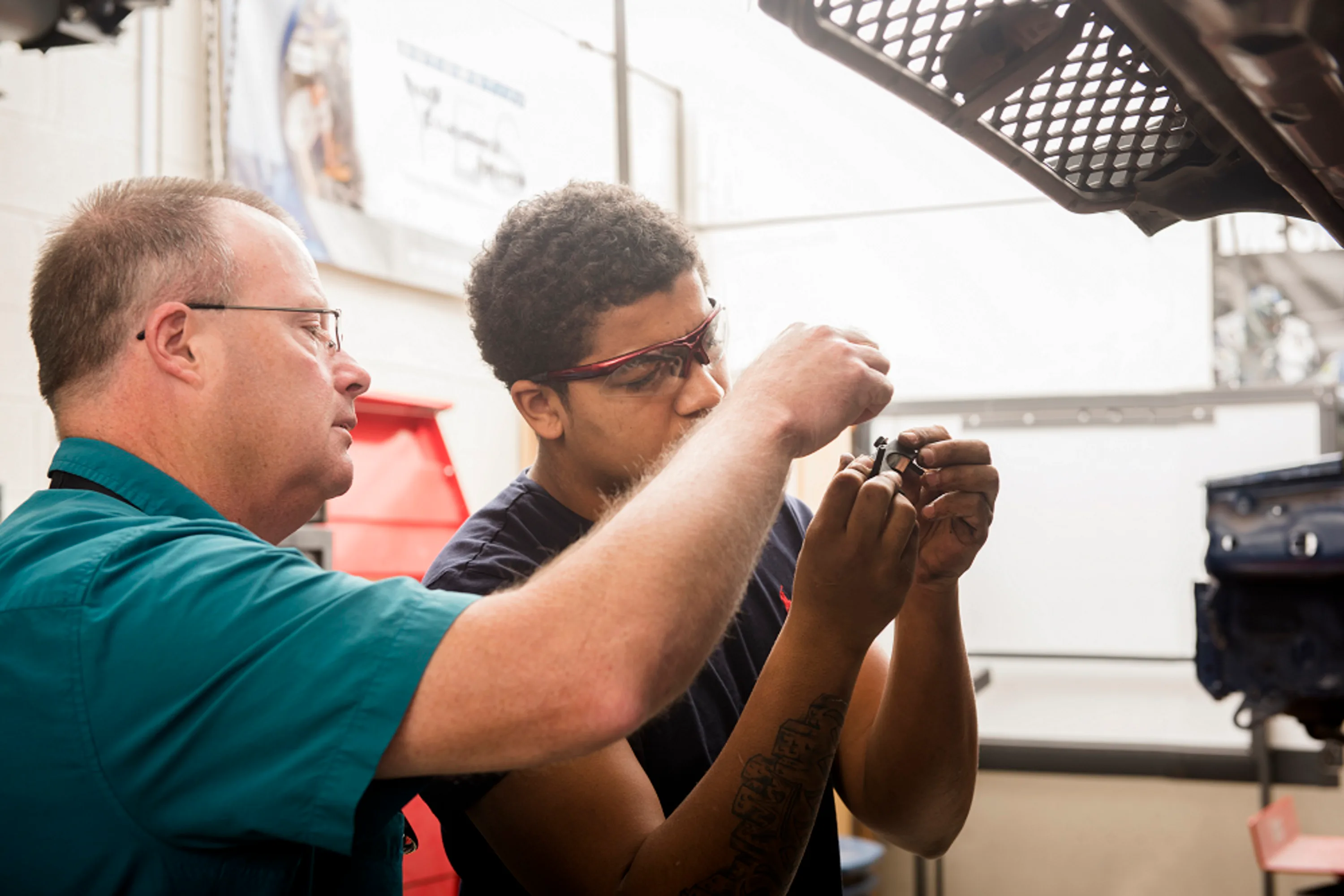 Instructor guiding a student working on a mechanical part in a shop