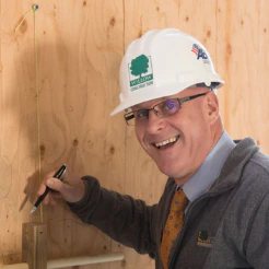 Smiling man in a white hard hat marking a measurement on plywood
