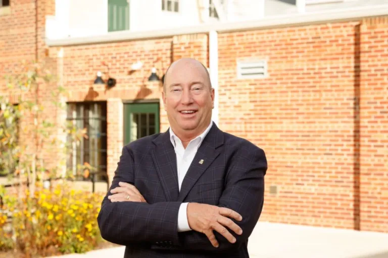 Smiling man in a dark blazer with arms crossed in front of a brick building