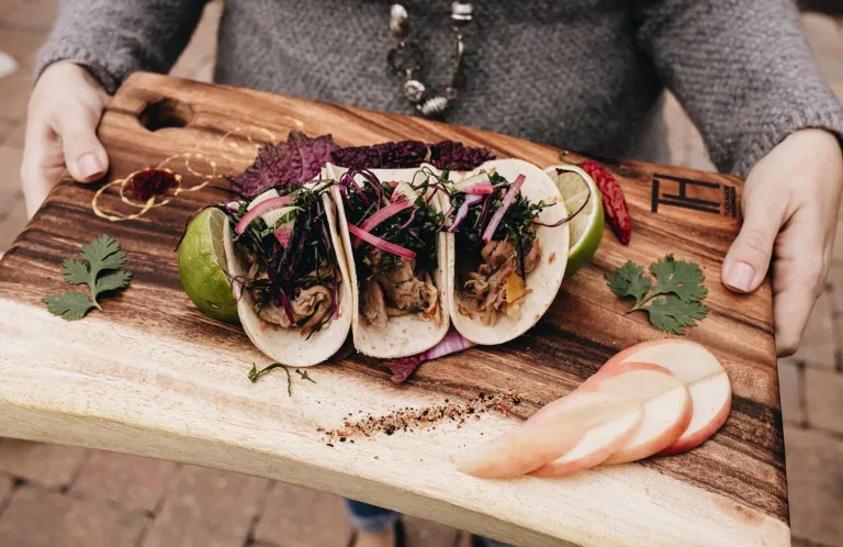 Hands holding a wooden board with three tacos garnished with herbs and onions
