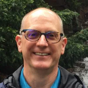 Smiling man with glasses and a blue shirt in front of evergreen trees