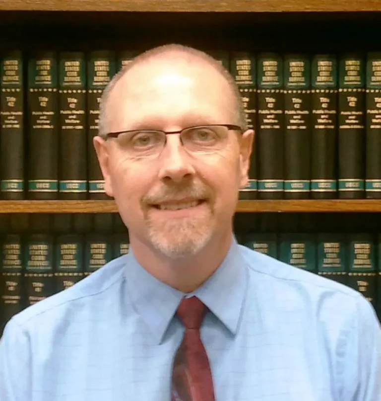 Headshot of a man in glasses and a blue shirt in front of a shelf of law books