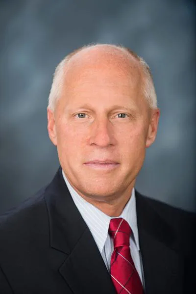 Headshot of a bald man in a dark suit and red tie against a gray background