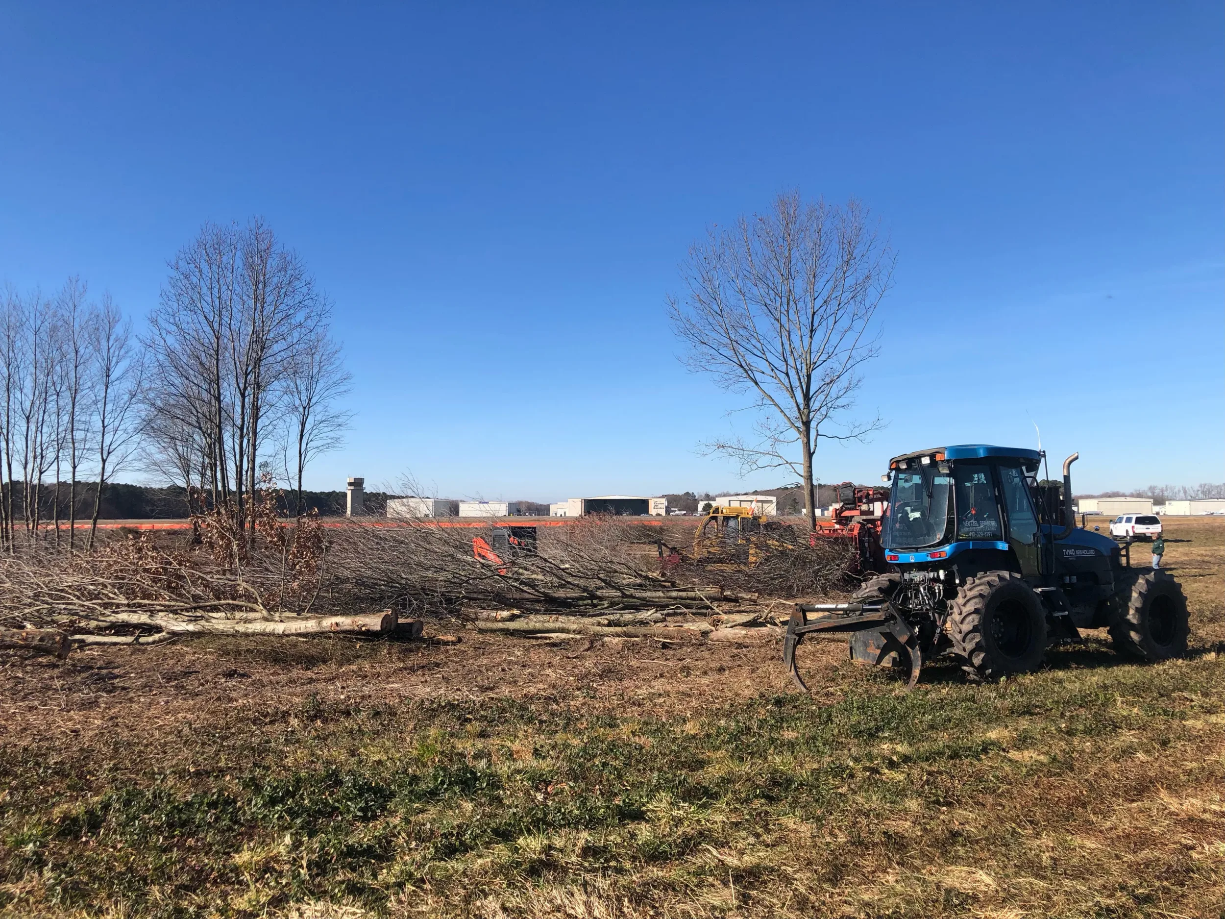 Blue tractor clearing trees from a field with cut logs piled nearby