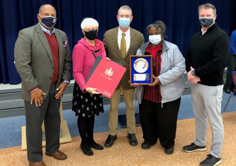 Five people posing with a plaque and a red folder in an auditorium