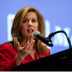 Woman in a red jacket gesturing while speaking at a microphone
