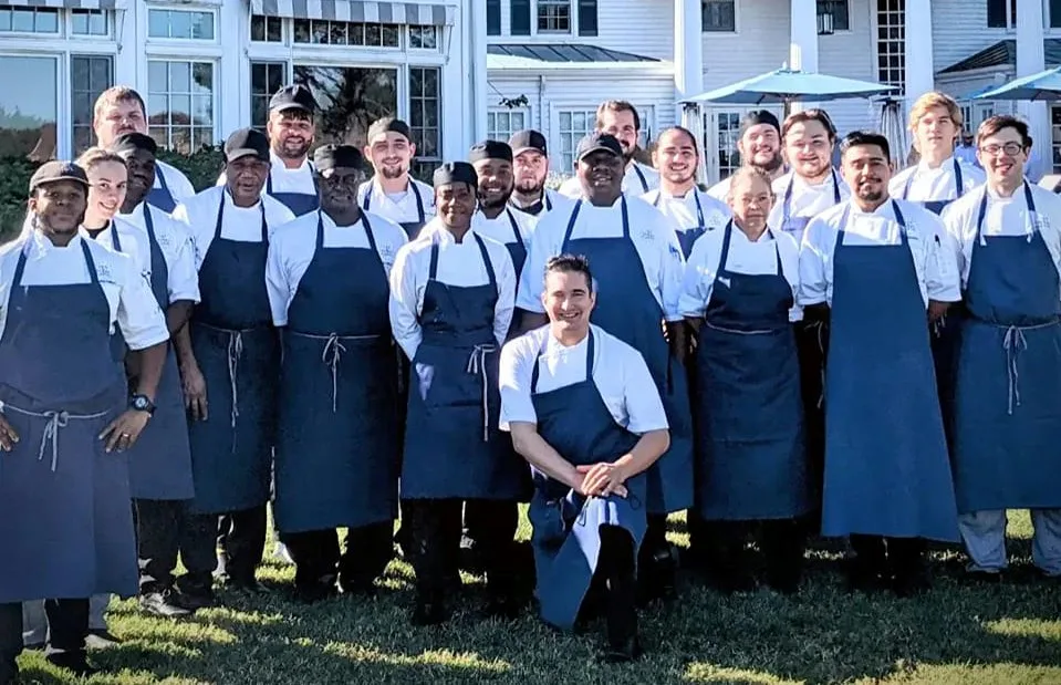 Large group of chefs in navy aprons standing outdoors in front of a white building