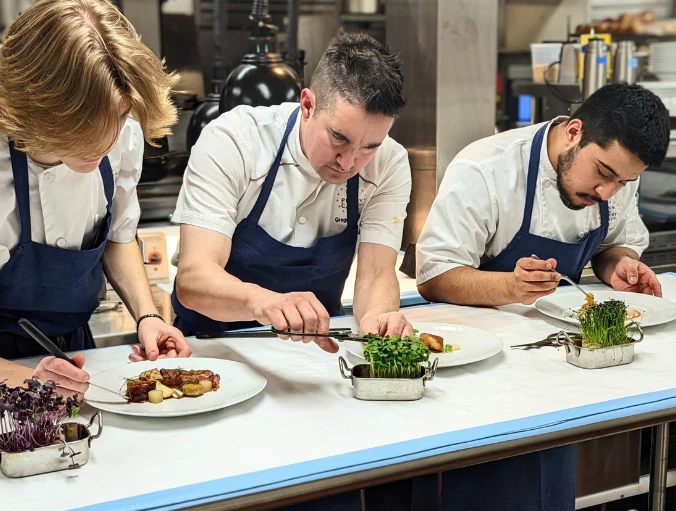 Three chefs in white coats plating dishes at a kitchen counter
