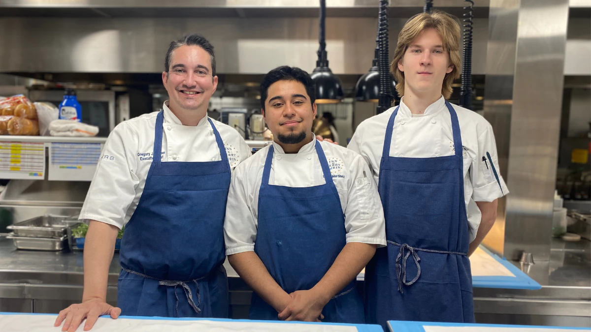 Three chefs in navy aprons and white coats standing in a commercial kitchen