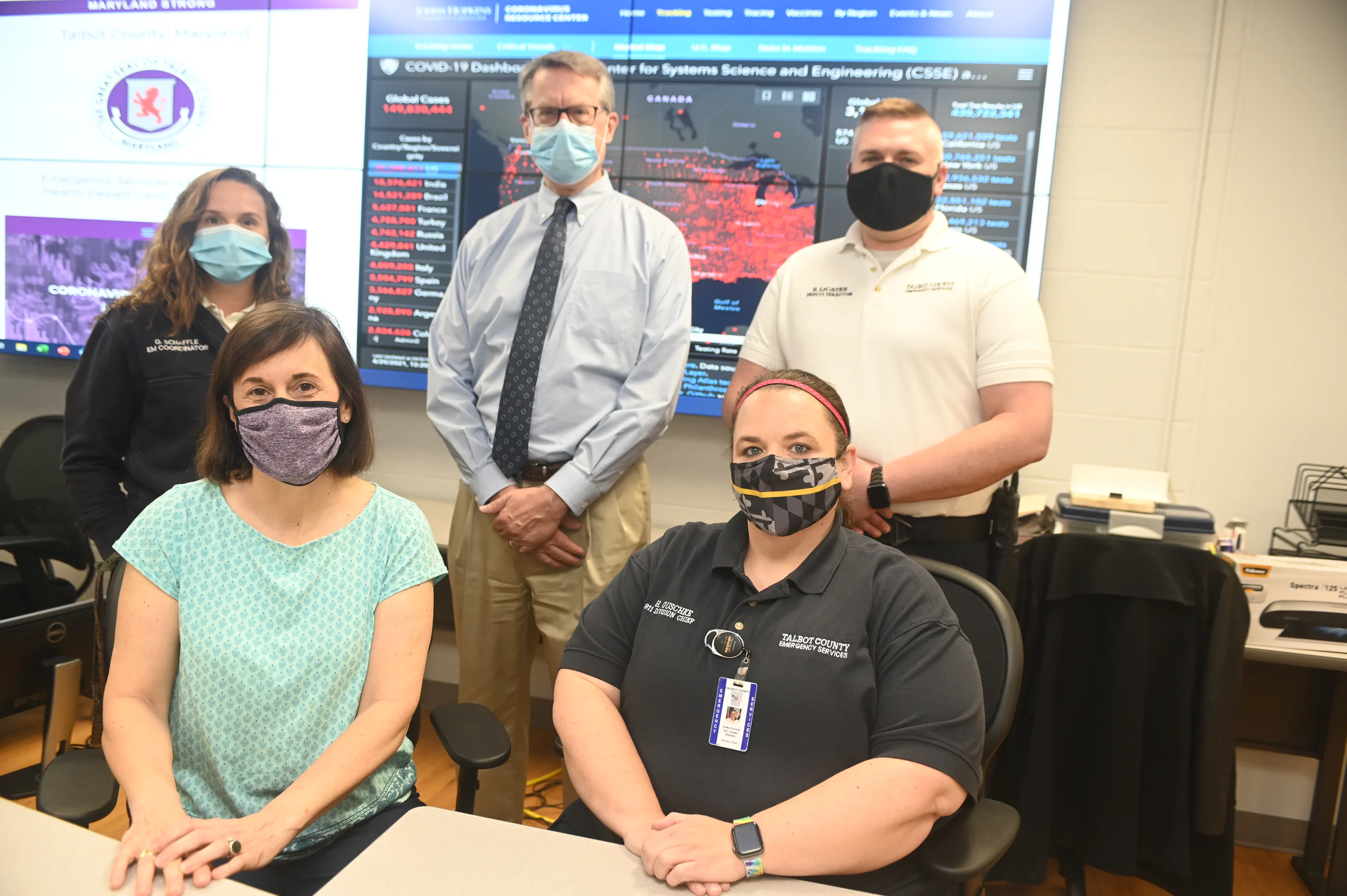 Five people seated and standing in a control room with monitors, wearing masks