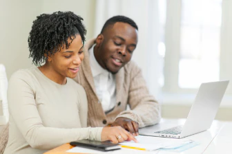 Man and woman smiling together at a laptop, reviewing papers at a table