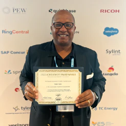 Man in dark suit holding an achievement award certificate in front of sponsor logos