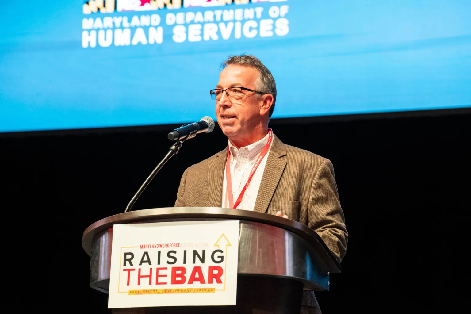 Man speaking at podium with Raising the Bar sign and Maryland Department of Human Services backdrop