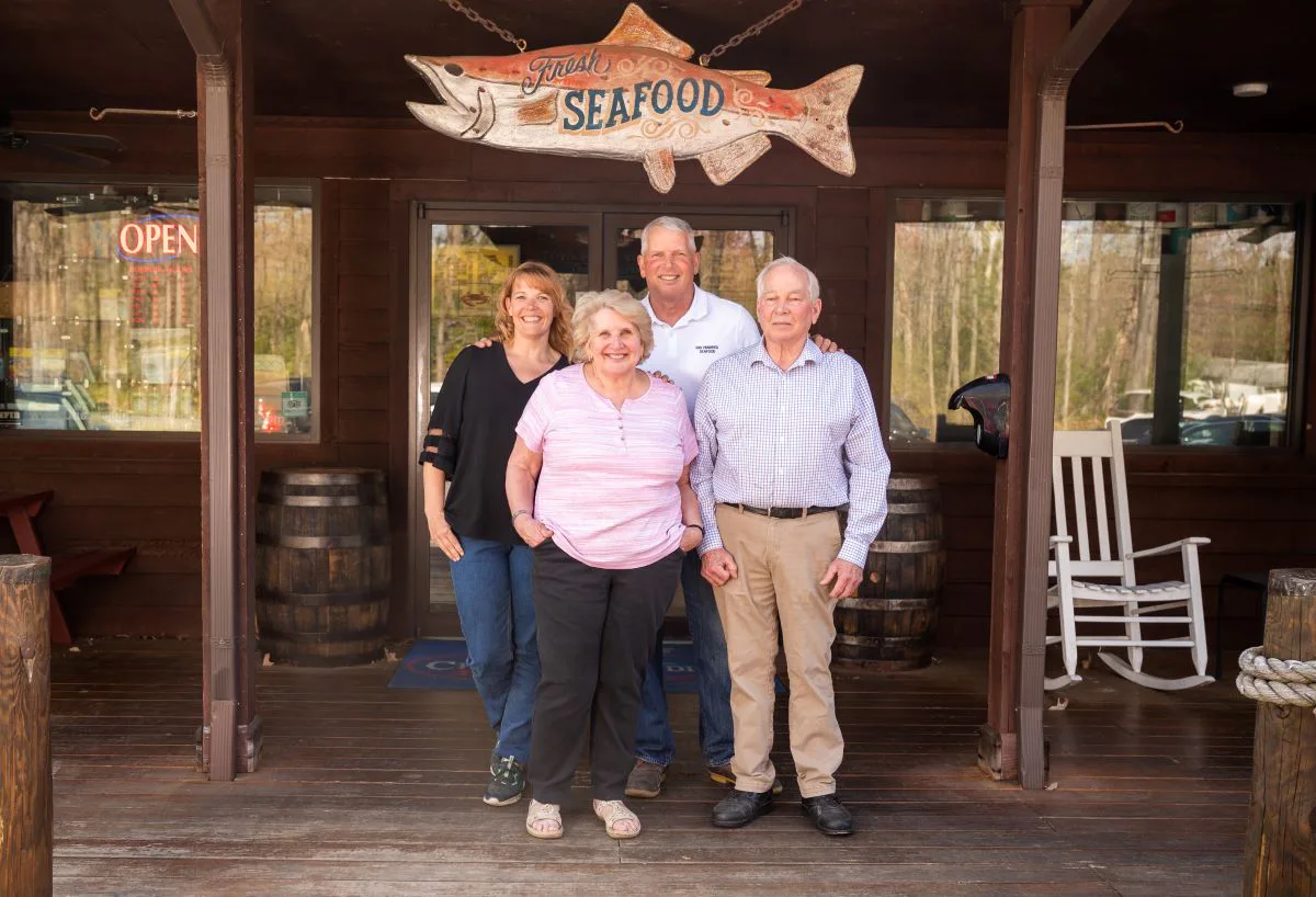 Four people posing in front of a rustic seafood sign with a fish cutout