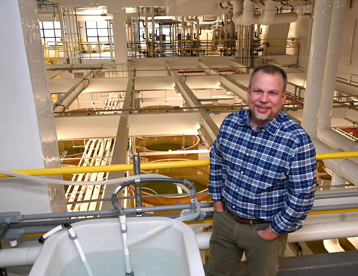 Man in blue plaid shirt standing among industrial pipes and tanks in a facility