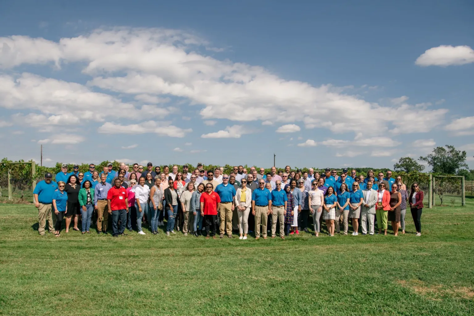 Large group of people posing on grass in front of a vineyard