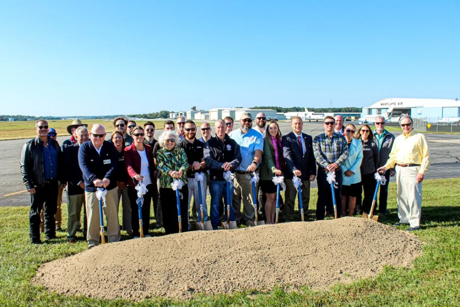 Group of about eighteen people with ceremonial shovels behind a dirt mound at an airport