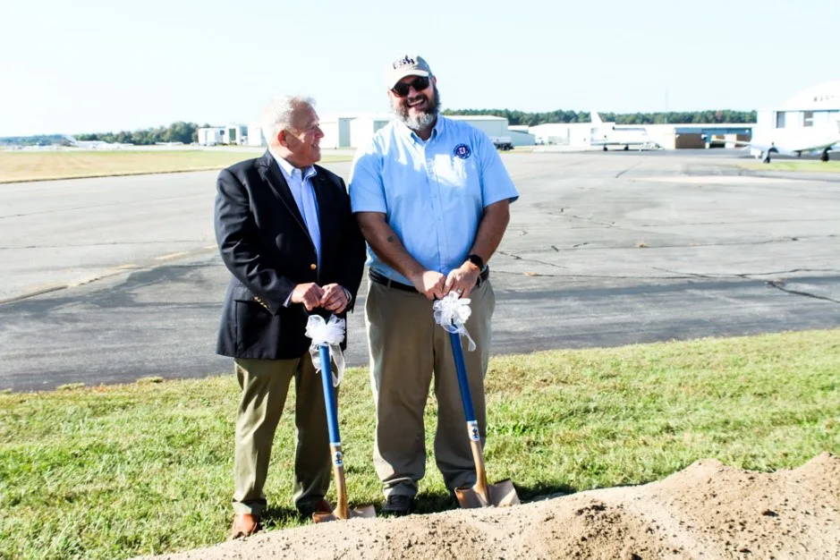 Two men holding ceremonial shovels beside a dirt mound on an airfield