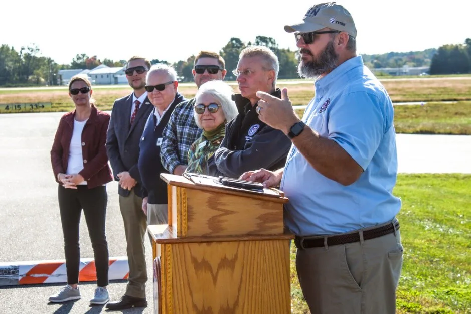 Officials speaking at a lectern on an airfield tarmac in front of a group