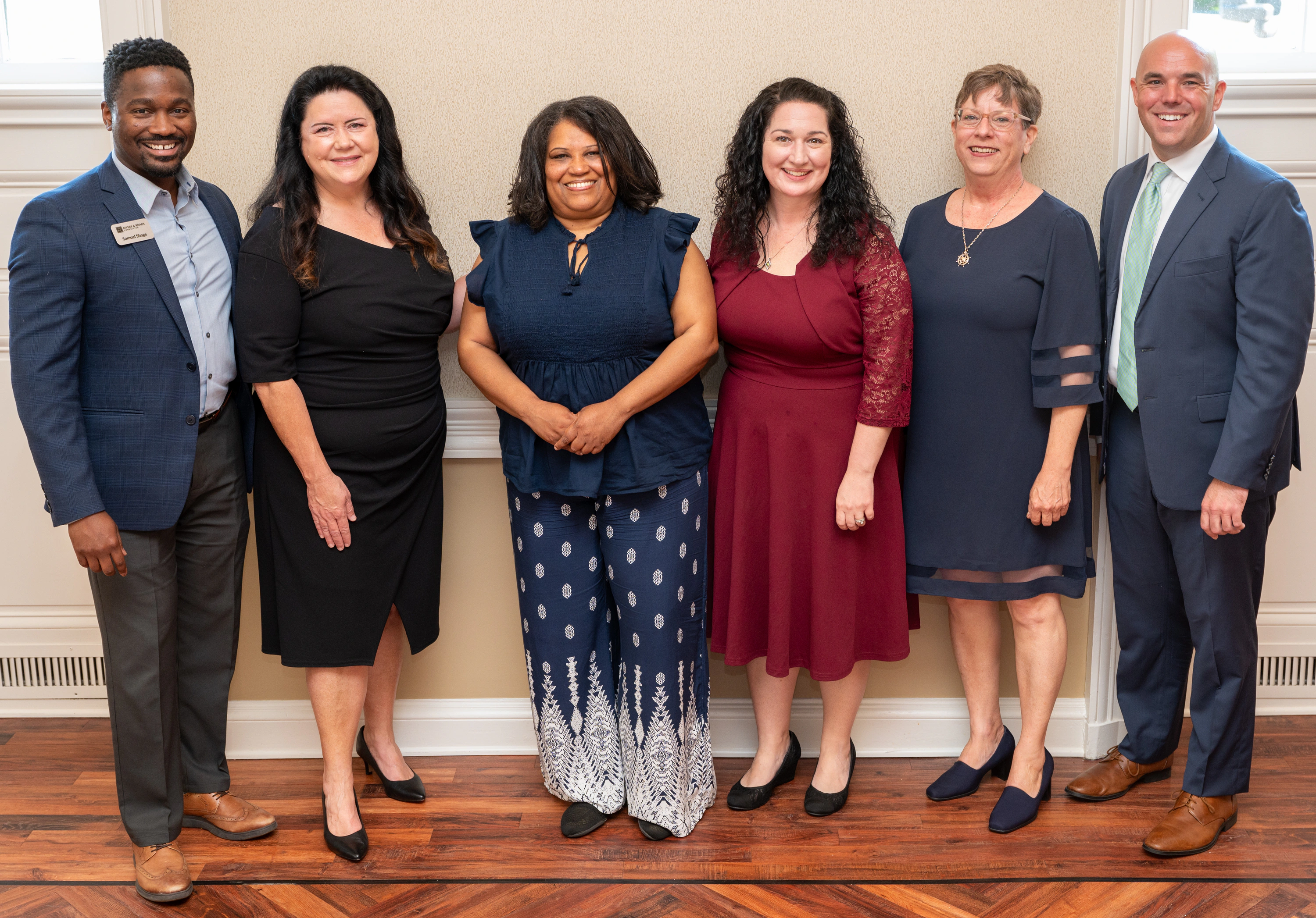 Six people in formal wear posing in a row against a wall
