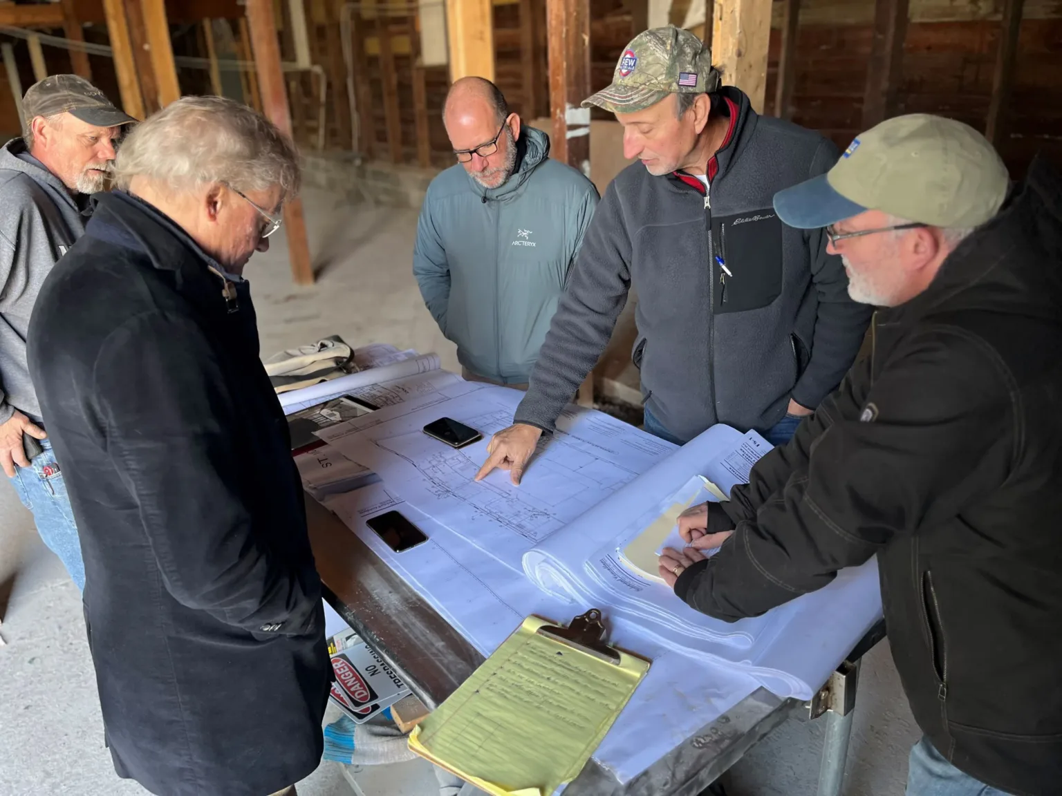 Five men gathered around a table of building plans in an unfinished wood-frame room