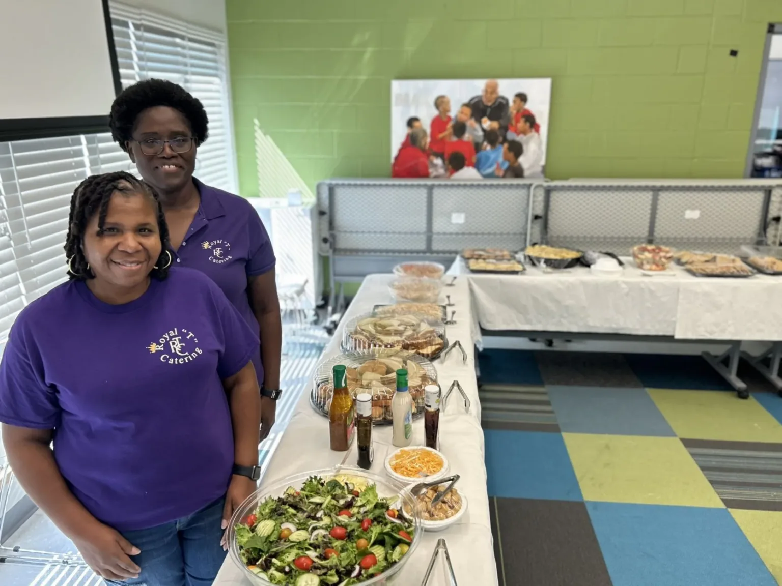Two women in purple shirts behind a salad and food table at an event