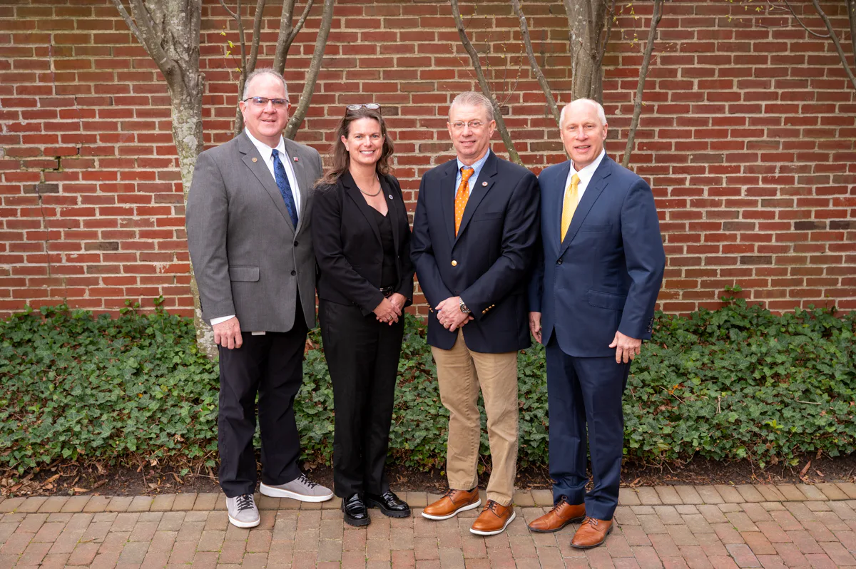 Four men and women in business attire stand together in front of a brick wall