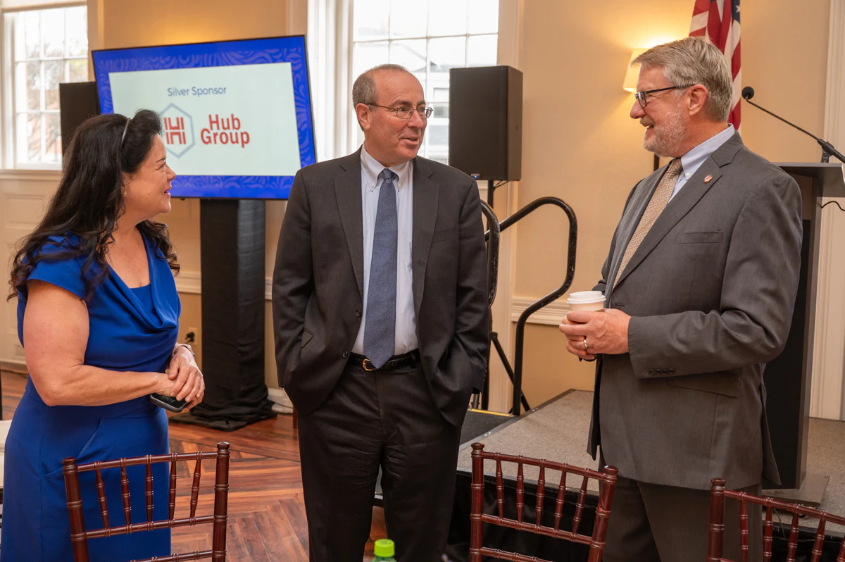 Woman in blue dress and two men in suits stand beside a screen at an event